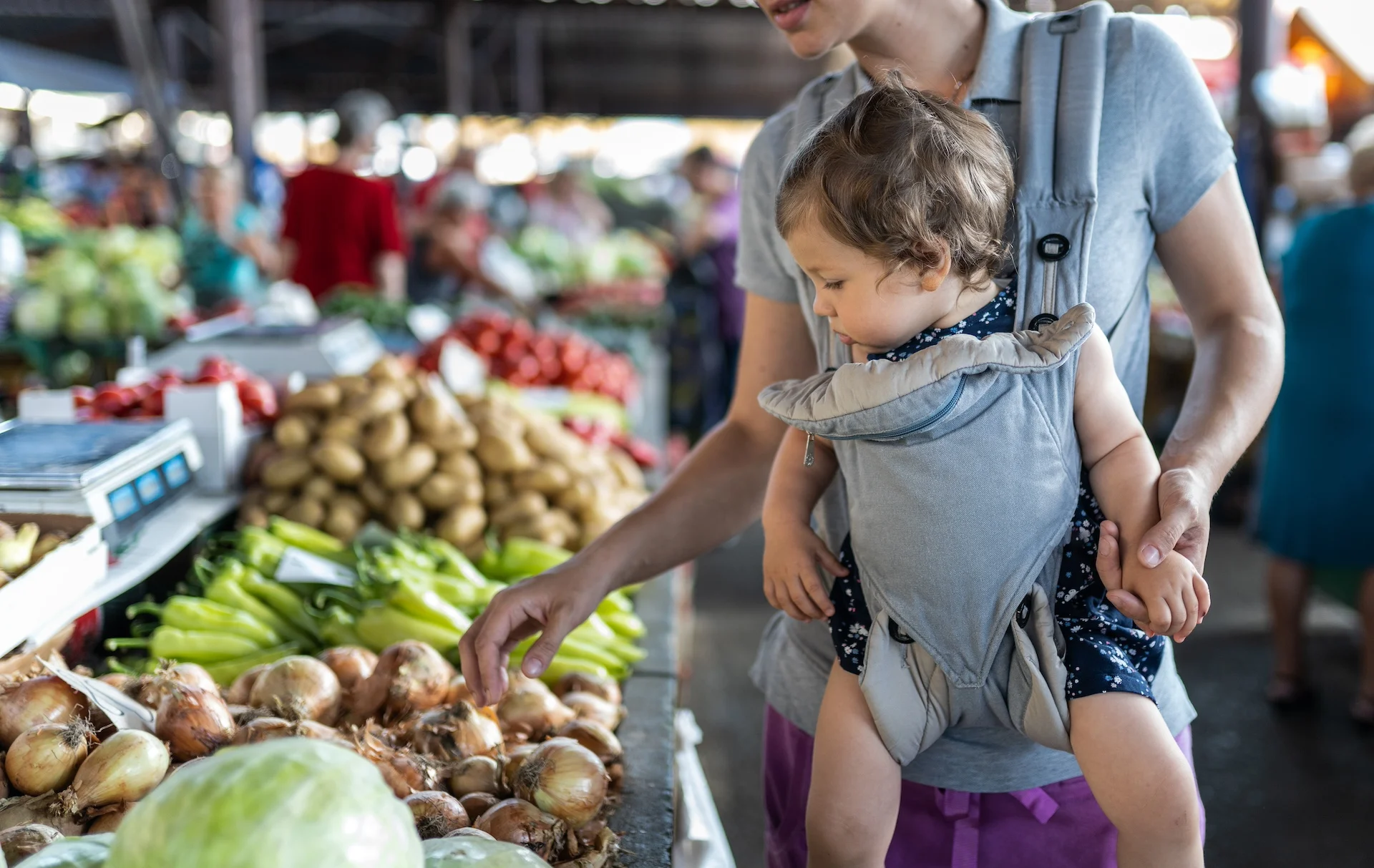 Mother-carrying-her-girl-in-baby-carrier-and-looking-vegetables-on-farmers-market